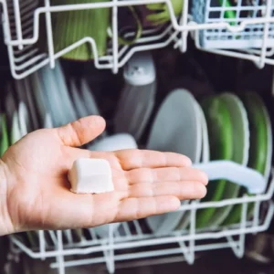woman's hand holding a homemade diy dishwasher tablet in front of a dishwasher