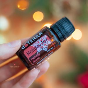 woman's hand holding a bottle of doterra holiday joy essential oil with sparkly lights in the background