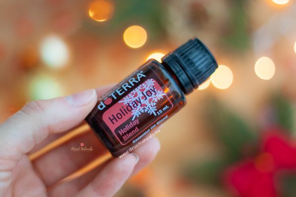woman's hand holding a bottle of doterra holiday joy essential oil with sparkly lights in the background
