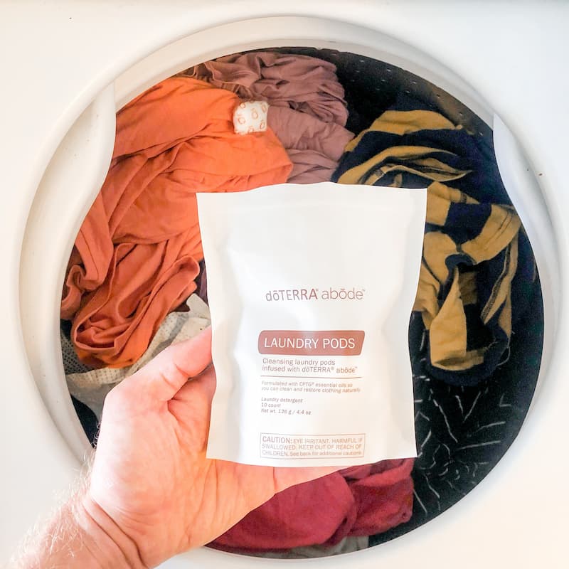 image of mans hand holding bag of doterra abode laundry pods in front of a front loading washing machine