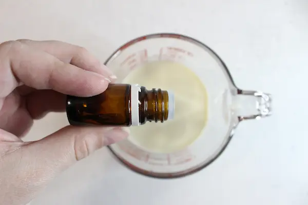 glass measuring cup with homemade sunscreen and a hand adding essential oil drops to it