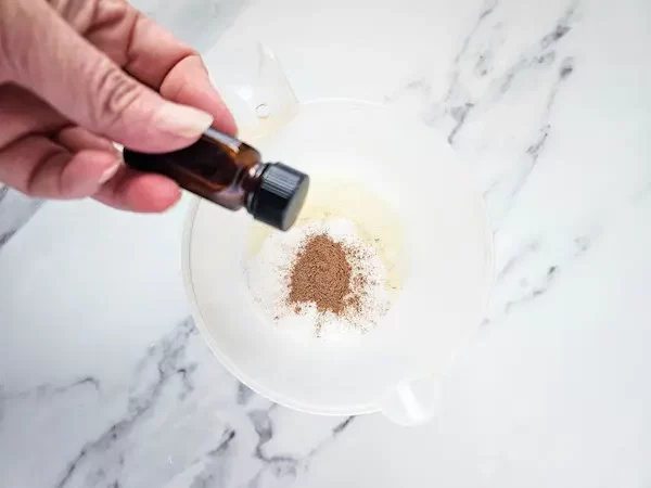 woman's hand holding bottle of essential oil over measuring cup filled with ingredients to make sugar scrub cubes