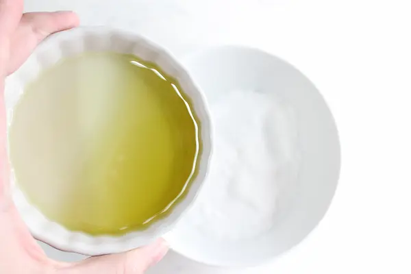 a hand holding a bowl of essential oil / carrier oil over a bowl of epsom salt and baking soda