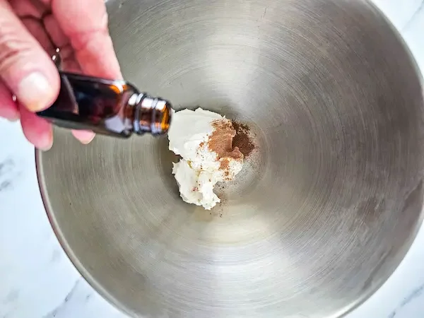 mixer bowl with homemade body butter in it and a hand adding drops from an essential oil bottle