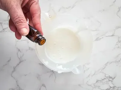 a woman's hand holding a bottle of essential oil and adding drops to a measuring cup filled with a melt and pour soap base mixture