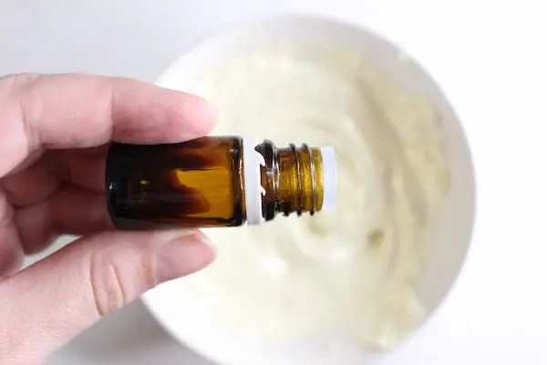 a woman's hand holding a bottle of essential oil over a bowl of homemade diaper rash cream