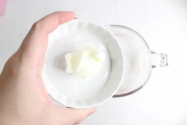 hand holding a small bowl of tallow, pouring it into a glass measuring cup filled with melted melt and pour soap base
