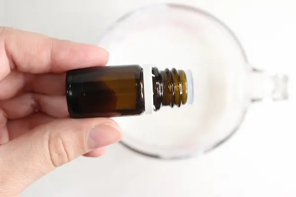 a hand holding a bottle of essential oil pouring drops into a glass measuring cup filled with melted soap base and tallow for making soap