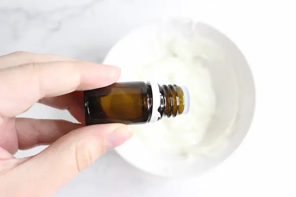 a hand holding a bottle of essential oil, adding drops to homemade whipped tallow balm in a bowl.