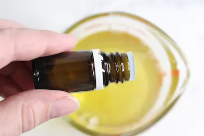glass jar of melted tallow with rose infused oil and a hand adding a few drops of essential oil to it