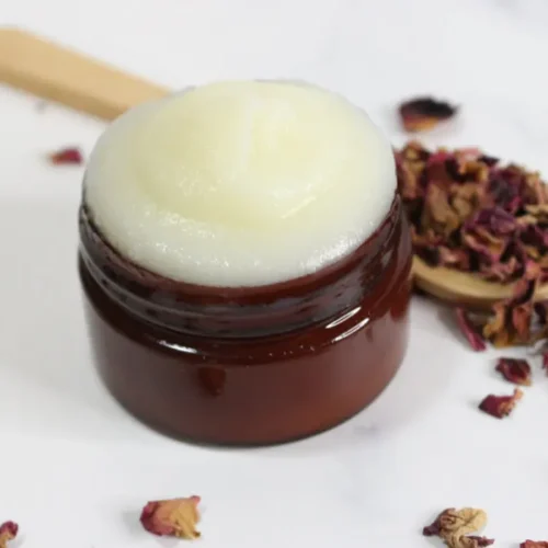 a jar of homemade rose tallow face cream with dried rose petals and a wooden spoon