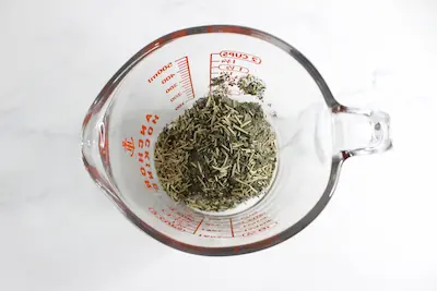 glass measuring cup with dried herbs in it for making rosemary lemon balm tallow stick