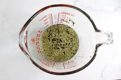 glass measuring cup with dried herbs and melted tallow in it for making rosemary lemon balm tallow stick