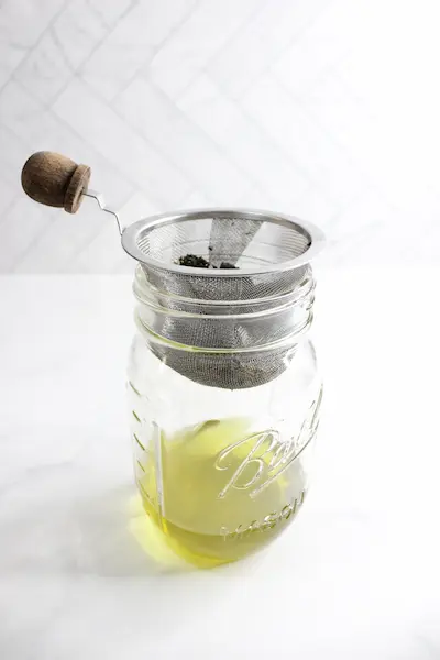 glass jar with melted tallow and a strainer on top with dried rosemary and lemon balm herbs