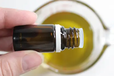 jar of melted beeswax, shea butter and tallow and a hand holding a bottle of essential oil adding drops to the mixture