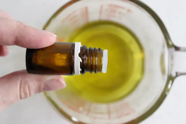 glass measuring cup with melted oils and butters in it with a hand dropping essential oil into for making a homemade sleep salve