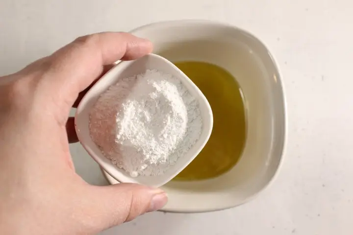 a woman's hand holding a bowl of zinc oxide powder over a bowl of oils to make diy sunscreen spray