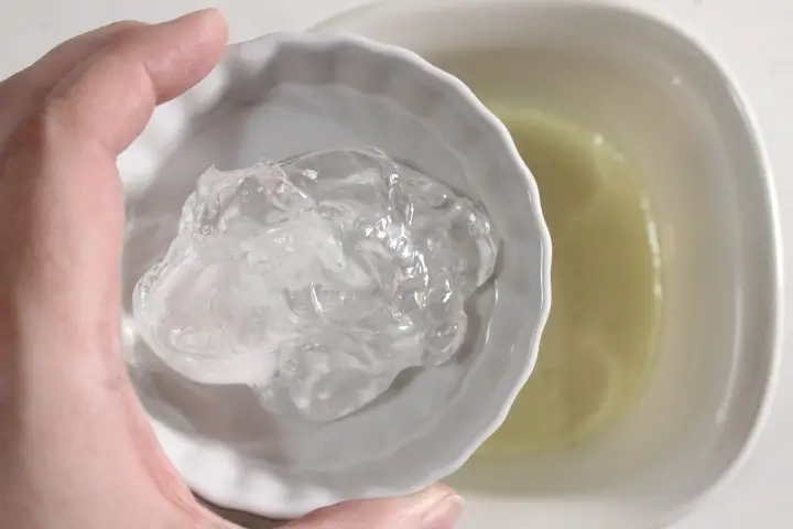 a woman's hand holding a bowl of aloe vera gel over a bowl of oils and zinc to make homemade sunscreen spray
