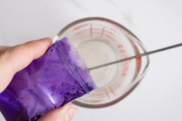 a pyrex measuring cup containing melted soap base and a woman's hand adding purple mica powder to the mixture for making easter bunny soaps